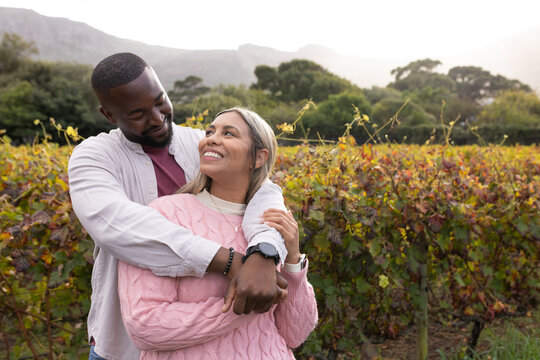 Diverse couple hugging and smiling while standing among vines in vineyard with watch, casual layers