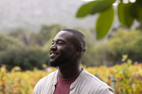 African man standing, smiling and looking left among vine rows with stakes, wearing maroon tee