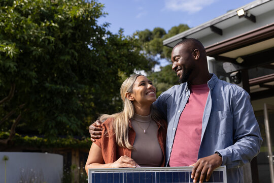 Couple holding rectangular solar panel at suburban home porch under mature trees, smiling warmly