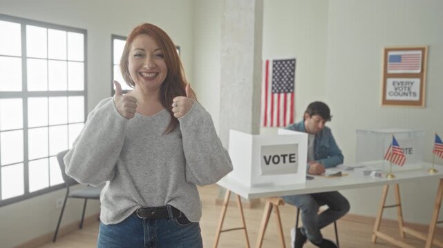Smiling woman giving thumbs up next to ballot box and voting booth in polling building while man signs forms at table; civic pride.