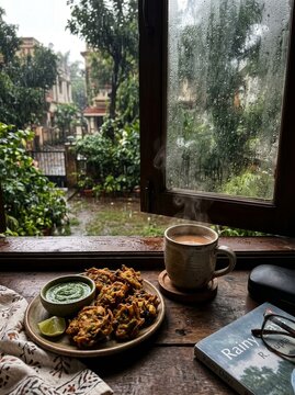 Cup of tea with pakora snacks on wooden windowsill during rain. Cozy home atmosphere with food and book overlooking rainy garden view. Relaxing afternoon break during monsoon season.