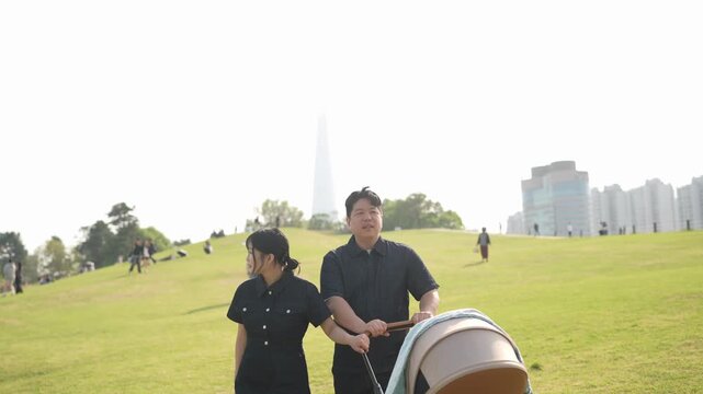 Soft light bathes a wide green hill. Man and woman walk side by side, pulling a stroller. Distant cityscape frames the open space. They look relaxed, enjoying a quiet outing