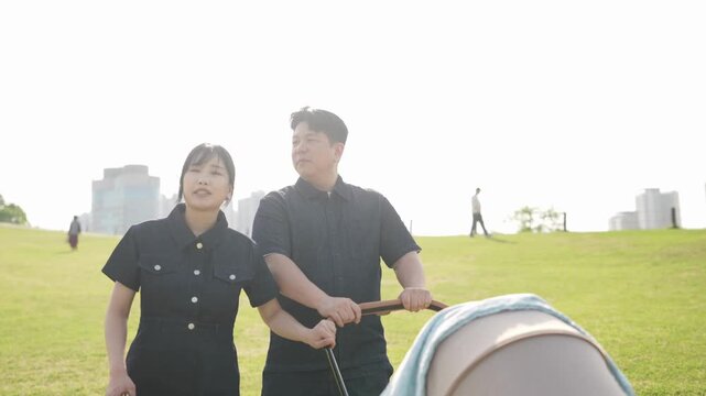 Soft light bathes a green hillside. Couple walks with a stroller. They share a quiet, contented moment. City buildings frame the distant background. The scene suggests peaceful family outings