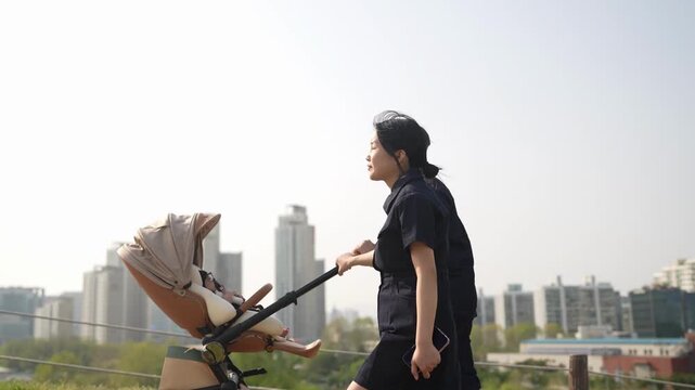 Woman pushes stroller on sunny rooftop. City skyline visible in distance. She smiles, pointing with confident gesture. Ideal for urban parenting or lifestyle themes. No animals or plants in scene