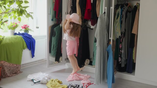 Carefree little girl wearing a huge adult pink sun hat jumps up at home.