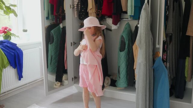 A lovely blonde girl in a pink hat tries on clothes by a wardrobe mirror.