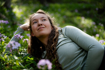 Fototapeta premium Young woman enjoying a peaceful moment among wildflowers in a green forest, lying on the meadow in sunlight, feeling joy, mindfulness, wellbeing and natural beauty.