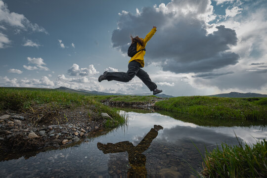 Active woman hiker with backpack jumping high in midair over mountain stream with water reflection. Female traveler exploring remote wilderness under dramatic cloudy sky.
