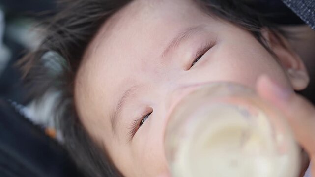Soft light catches a baby's peaceful face. Closed eyes show quiet contentment as they drink. Gentle hand holds the bottle steady. Perfect for scenes of calm, tender parenting moments