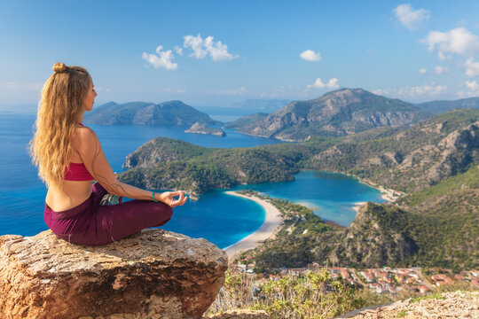Young woman sit in yoga pose on rock top relaxing, looking at blue lagoon, Oludeniz beach. Summer day walk by Lycian way at family vacation in Mediterranean Sea coast, Fethye, Antalya region, Turkey