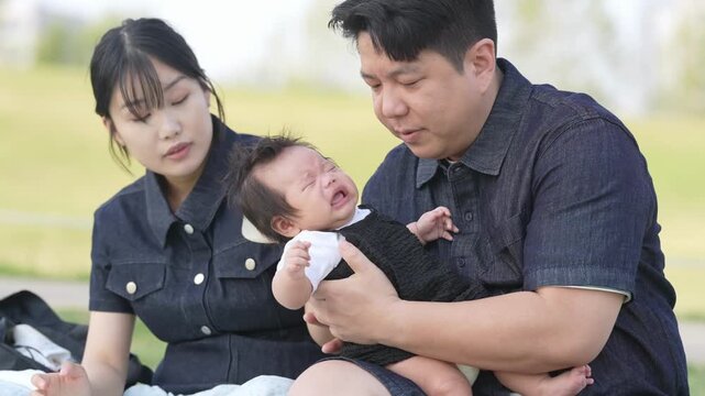 Baby cries loudly while parents sit on grass. Mother holds bottle, father holds baby. Parents look tired and concerned. Perfect for family life, parenting, or childcare scenes