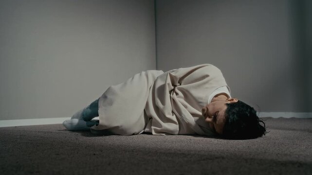 Eye level shot of adult woman wearing straitjacket lying in fetal position on floor of psychiatric clinic. Video is ideal for mental health awareness campaigns and medical documentaries