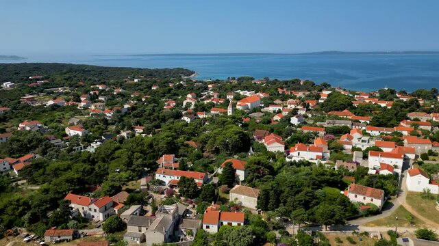 An aerial drone shot overlooking the charming, sun-drenched village on Silba Island, Croatia. The perspective captures traditional stone houses with bright red roofs nestled within lush green