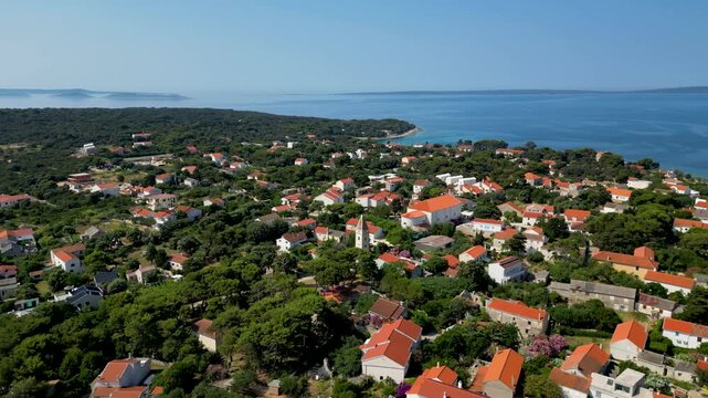 An aerial drone shot overlooking the charming, sun-drenched village on Silba Island, Croatia. The perspective captures traditional stone houses with bright red roofs nestled within lush green