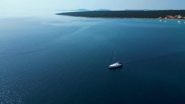 A stunning aerial drone perspective showcasing a white sailboat gliding through the crystal-clear, turquoise waters of Silba Island, Croatia. The shot beautifully captures the contrast between the