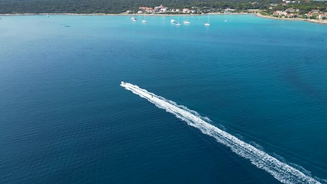 An exhilarating aerial drone shot over Silba Island, Croatia, capturing a water skier carving through the crystal-clear, turquoise sea behind a speeding motorboat. The top-down view beautifully frames