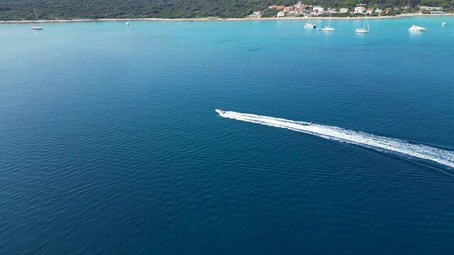 An exhilarating aerial drone shot over Silba Island, Croatia, capturing a water skier carving through the crystal-clear, turquoise sea behind a speeding motorboat. The top-down view beautifully frames