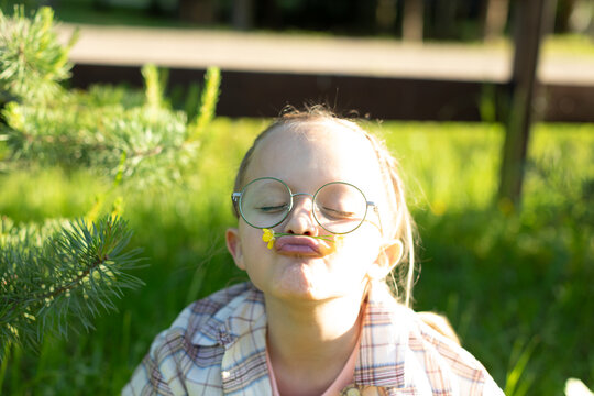 Funny little girl in round glasses holding a yellow flower with her lips and making a face in the sunny summer garden