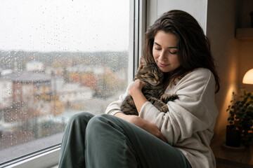 Fototapeta premium A woman sits by a window, holding her cat close while rain falls outside. The scene conveys warmth and comfort in a cozy indoor environment.