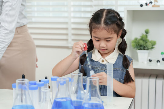 asian girl student sitting in classroom performing science experiment with dropper and plastic bottle focused on transferring blue solution into glass beaker among lab equipment in elementary school