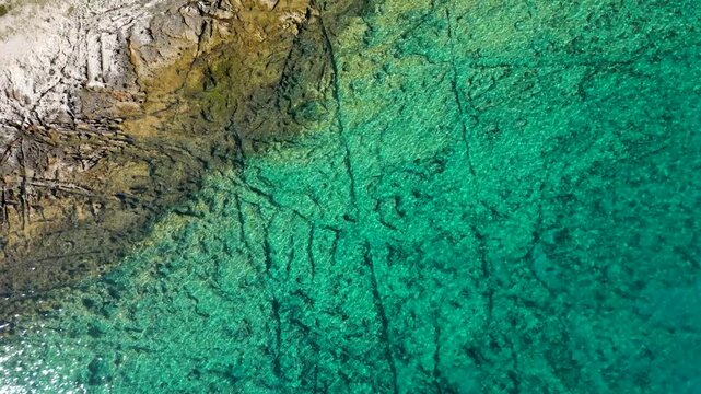 A stunning top-down aerial drone shot capturing the crystal-clear, emerald-turquoise waters of Silba Island, Croatia. The transparent sea beautifully reveals the rocky textures and patterns of the
