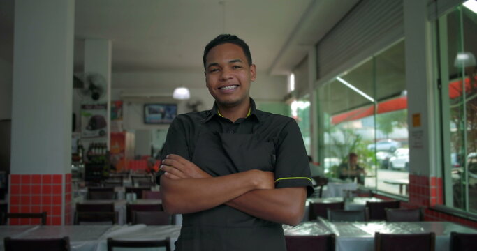 Latino restaurant employee smiling with arms crossed, cheerful and confident body language in bright indoor restaurant setting
