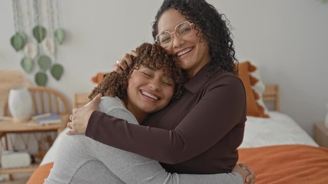 Woman hugging sister on bed in building, arms wrapped around shoulders, smiling with eyes closed, head resting near chest; comfort intimacy warmth.