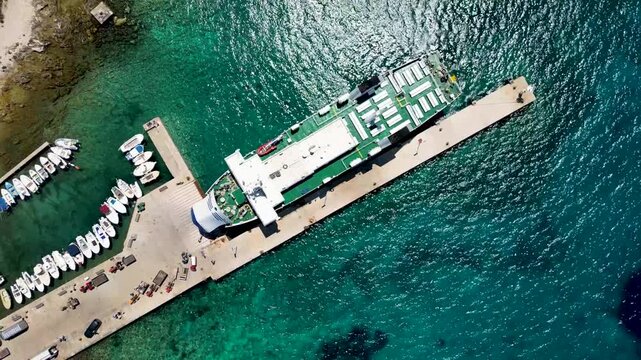 Top-down drone view of a ferry docked at Zalic port on Silba Island, Croatia. Crystal-clear Adriatic waters, marina facilities and moored boats highlight maritime transport, island connectivity and