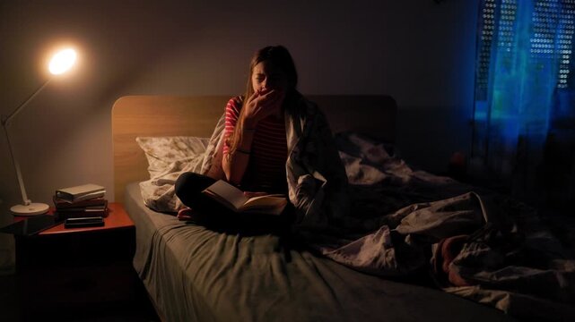 Tired preteen yawning and rubbing face while reading book in bed