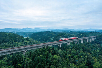 Obraz premium Red Train Traveling on Elevated Railway Bridge Over Dense Forest with Mountain Range in Background