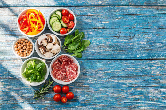 Fresh Ingredients Including Sliced Salami, Bell Peppers, Cherry Tomatoes, Cucumber, Mushrooms, Spinach, Basil, Chickpeas, and Herbs on Blue Wooden Table