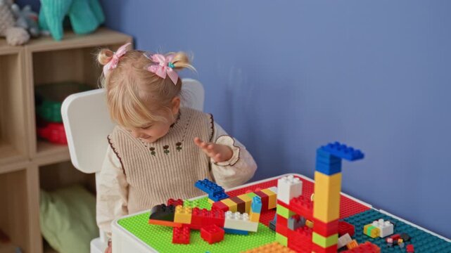 Little blonde girl reaches into a toy bin while assembling colorful blocks on a table in a building; quiet concentration.