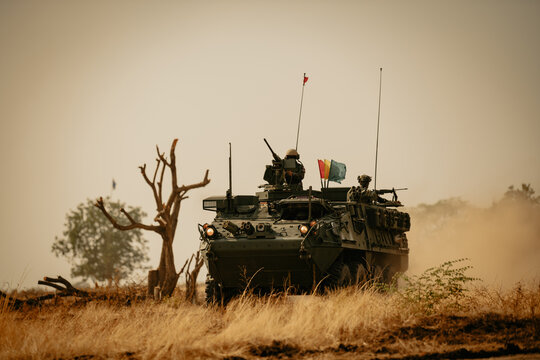 Dramatic shot of an eight-wheeled green military armored vehicle (APC) advancing through a dense dust and haze cloud on a dry dirt field under heavy golden sunset light.