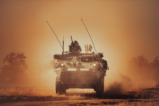 Dramatic shot of an eight-wheeled green military armored vehicle (APC) advancing through a dense dust and haze cloud on a dry dirt field under heavy golden sunset light.
