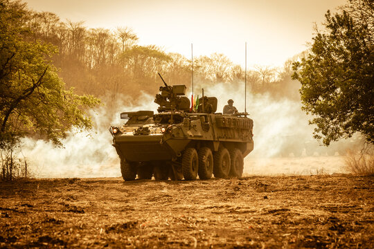 n eight-wheeled military armored personnel carrier (APC) moving fast on dry ground, kicking up dust and smoke under a dramatic golden orange sunset sky