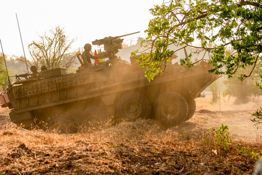 A powerful low-angle shot of a green armored military vehicle (APC) leaping over a dirt ridge, framed by tree branches under a brilliant golden sunset sky.