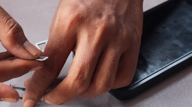 Close-up view of an Asian  man's hands carefully swapping sim card inside smartphone, sitting at table with grey surface with extraction tray nearby during mobile service transition.