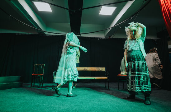 Young actors performing on a small theatre stage in period costumes and props. A rehearsal scene with dramatic green lighting and expressive poses from the cast.