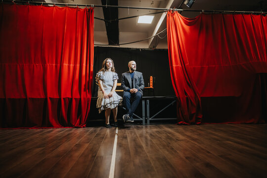 Two young actors sit at the front of a stage framed by red curtains. A male and female performer wait during a quiet theater rehearsal under stage lighting.