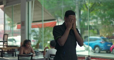Fototapeta premium Restaurant worker covering face with both hands near window, showing signs of burnout, emotional exhaustion, and overwhelming fatigue