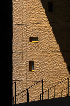 Textured yellow wall with small windows falls into deep shadow, where staircase railing, urban architecture and sunlight create strong contrast