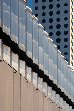 A glass facade and dotted tower rise beside beige wall panels, where windows, shadow, urban architecture and vertical geometry shape depth