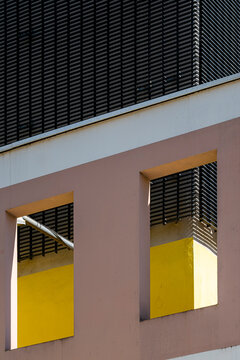 A pink building wall with yellow windows and black shutters uses sunlight, shadow and urban architecture to turn facade into color geometry