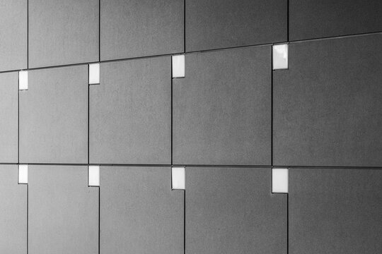 Grey wall panels create a precise grid across a concrete facade, where small squares, shadow and urban architecture become abstract pattern