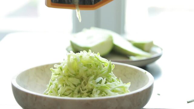 Close-up of a woman's hands grating a fresh green zucchini on a metal box grater in a bright kitchen