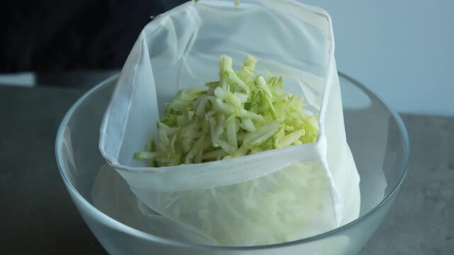 Close-up of a woman's hands squeezing and draining liquid from grated zucchini inside a mesh bag
