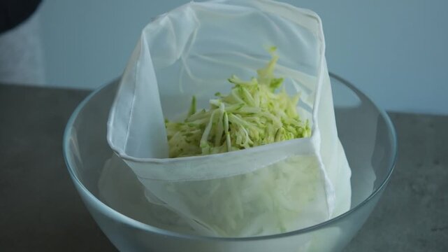 Close-up of a woman's hands squeezing and draining liquid from grated zucchini inside a mesh bag