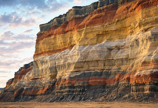Close up of a steep cliff face showing colorful sedimentary rock layers in shades of red, yellow, and white under a soft evening sky