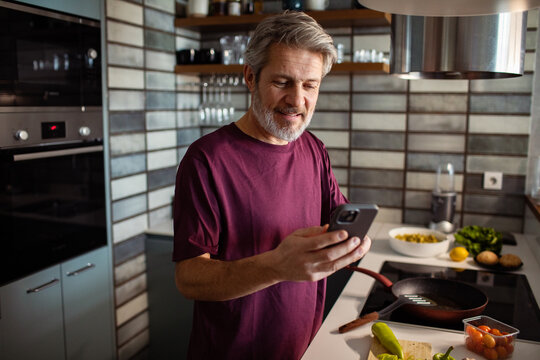Middle aged man checking recipe on smartphone while cooking in modern kitchen