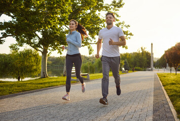 Fototapeta premium Smiling athletic young running together in park on sunny day. Energetic sporty man and woman in sportswear jogging during outdoor workout and staying active in healthy lifestyle routine